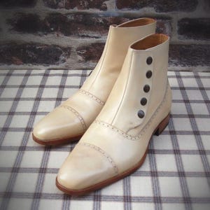 Puede incluir: Un par de botines de cuero color crema. Los botines tienen cierre de botones en el lateral, punta redondeada y suela marrón. Costuras decorativas acentúan la puntera y los laterales. Los botines se exhiben sobre una superficie a cuadros.