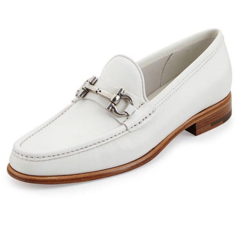 May include: White leather loafer with a silver-tone metal buckle detail. The shoe has a rounded toe, a low heel, and a stitched design around the edges. The sole is a light brown color.