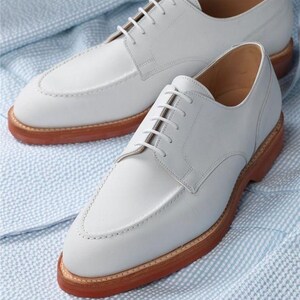 May include: A pair of white leather dress shoes with a classic design. The shoes have a light gray hue, a stitched upper, and brown soles. The shoes are displayed on a light blue and white patterned fabric.