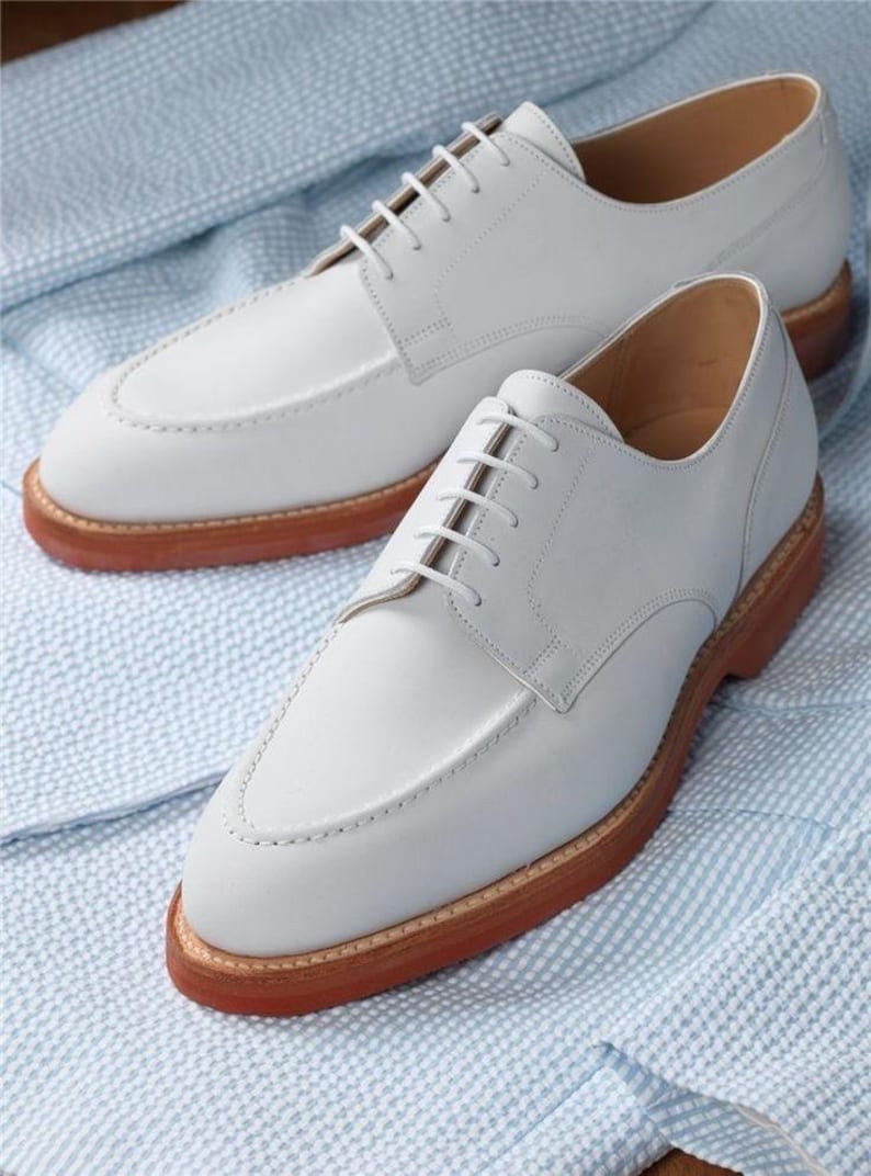 May include: A pair of white leather dress shoes with a classic design. The shoes have a light gray hue, a stitched upper, and brown soles. The shoes are displayed on a light blue and white patterned fabric.