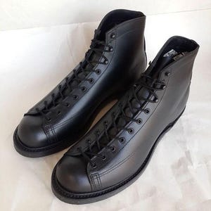 Puede incluir: Un par de botas de cuero negro con diseño de cordones. Las botas tienen punta redonda y un estilo de caña alta. Las suelas son negras y las botas están sobre un fondo blanco.