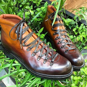 May include: A pair of brown leather hiking boots with dark gray laces. The boots have a high-top design with metal eyelets and a black sole. The boots are displayed on a bed of green plants.