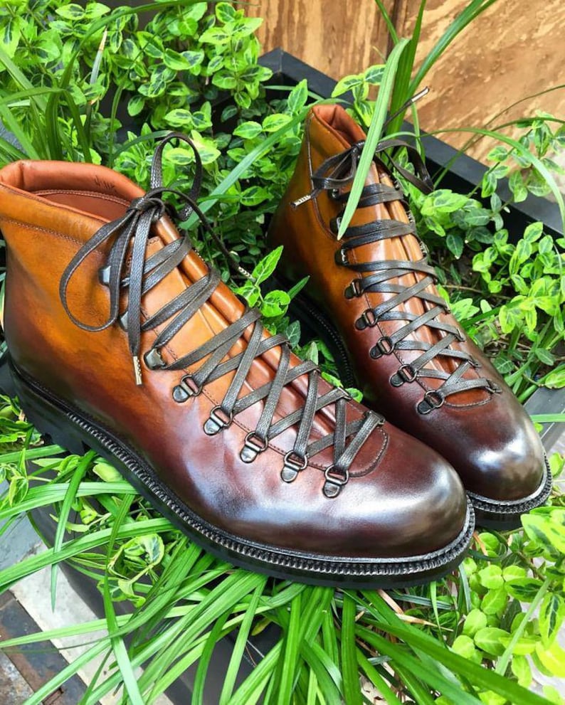 May include: A pair of brown leather hiking boots with dark gray laces. The boots have a high-top design with metal eyelets and a black sole. The boots are displayed on a bed of green plants.