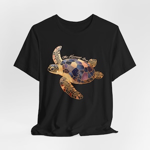 May include: A black short-sleeve t-shirt featuring a graphic of a sea turtle with a shell patterned in shades of blue, purple, and orange, accented with gold lines. The text "My Kintsugi Era" is printed in a light script font above the turtle.