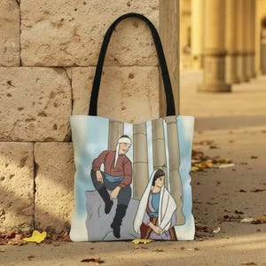 Pode incluir: Bolsa tote com alça preta. A bolsa exibe uma ilustração de duas figuras em um cenário clássico. Uma figura está sentada em uma rocha e a outra está sentada no chão. O fundo inclui colunas e um céu azul claro.