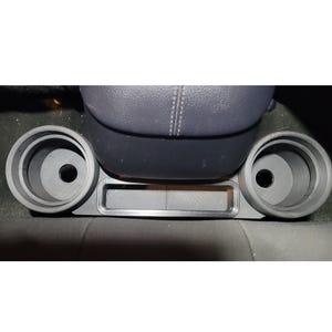 May include: A gray plastic car console with two circular cup holders and a rectangular storage area. The console is positioned below a dark gray car seat.