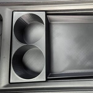 Könnte beinhalten: Ein schwarzer Kunststoff-Auto-Konsolen-Organizer mit zwei kreisförmigen Getränkehaltern und einer rechteckigen Ablage. Der Organizer ist für den Einbau in die Mittelkonsole eines Autos konzipiert und bietet Stauraum und Organisation.
