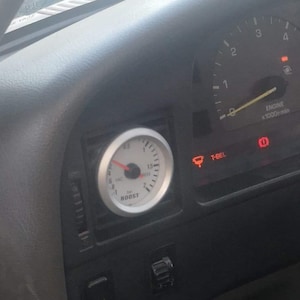 Puede incluir: Primer plano del salpicadero de un coche con un manómetro de sobrealimentación, un velocímetro y otros indicadores. Los indicadores están iluminados en rojo. El salpicadero del coche es negro y gris.