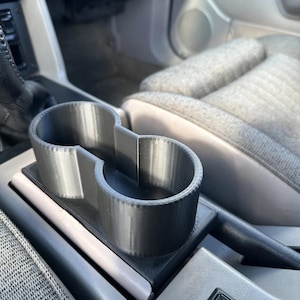 May include: A 3D-printed, dual-cup holder in a car's center console. The holder is gray and designed to fit into the existing console space. The car's interior features gray fabric seats and a black control panel.