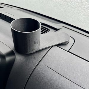 May include: A black car cup holder with a flat base, designed to sit on a car's dashboard. The cup holder is cylindrical and made of a matte black material. The dashboard is also black and has a textured surface.