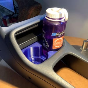 May include: A dark gray car console with a built-in cup holder and a small storage compartment. A purple water bottle with a white lid sits in the cup holder. The bottle has a label that reads "na'gene" and "SPOOL3D".