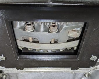 May include: A black rectangular automotive part with rounded corners, secured by screws, frames a metallic engine component. The engine part has bolts and a curved, toothed belt. The image is a close-up shot.