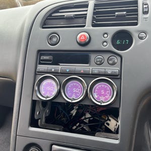 Könnte beinhalten: Autoinstrumententafel mit drei runden Messgeräten, die Temperaturwerte anzeigen. Die Messgeräte haben violette Zifferblätter und weiße Zahlen. Das Armaturenbrett ist grau mit verschiedenen Knöpfen und einer Digitaluhr, die 6:17 anzeigt.