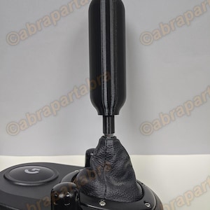 Può includere: Pomello del cambio nero con soffietto in pelle nera. Il pomello ha una superficie testurizzata e una piccola vite nella parte inferiore. Il soffietto è fissato a una base nera con un pulsante circolare.