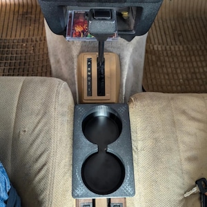 May include: A black, 3D-printed car cup holder with two circular openings. The cup holder is positioned between two beige car seats. The car's gear stick and dashboard are visible in the background.