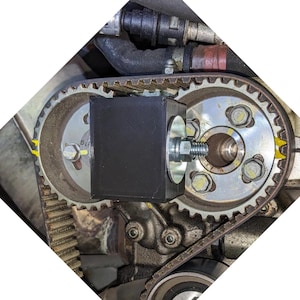 Peut inclure: Gros plan sur un composant de moteur avec un bloc noir rectangulaire fixé avec un boulon et une rondelle. L'ensemble est positionné entre deux engrenages métalliques reliés par une courroie dentée, avec des marquages jaunes.