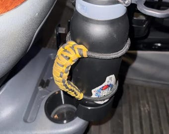 May include: A black water bottle with a yellow and black braided handle, secured in a car's cup holder. The bottle has a silver lid and a sticker. The handle is attached to the bottle with a black elastic cord.