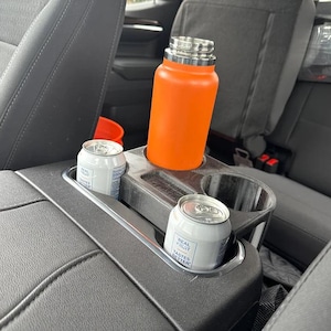 Puede incluir: Una botella de agua naranja aislada y dos latas de refresco Real Fruit están en los portavasos de la consola central de un coche. La consola es negra y tiene un borde plateado. Los asientos del coche son grises.