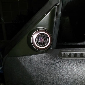 May include: A black car interior with a round speaker mounted in the door panel. The speaker has a black grille and a silver ring around the edge.