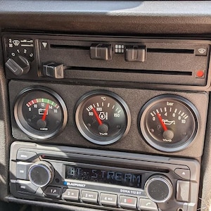 Può includere: Un cruscotto di un'auto nero con una radio, indicatori e un pannello di controllo. La radio sta riproducendo una stazione chiamata "BT STREAM" e gli indicatori mostrano la temperatura, la pressione dell'olio e la tensione della batteria dell'auto.