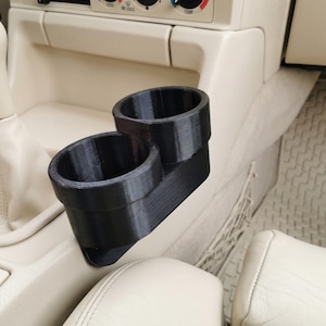 May include: Black dual cup holder for a car, designed to fit in the center console. The cup holder is made of a 3D-printed material and is shown in a car interior with a cream-colored dashboard and seats.