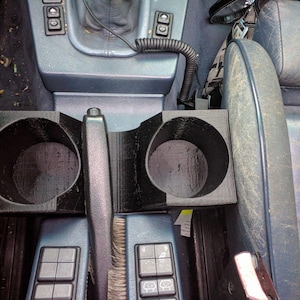 May include: Black 3D-printed car cup holder with two circular openings and a central divider. The cup holder is designed to fit in a car's centre console, near the gear shift and handbrake.