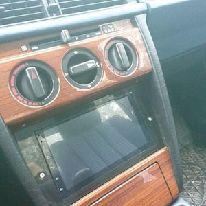 Puede incluir: Primer plano del salpicadero de un coche con acabado de madera. El salpicadero cuenta con una pantalla táctil, tres botones redondos y una rejilla de ventilación rectangular. La pantalla muestra una pantalla negra.