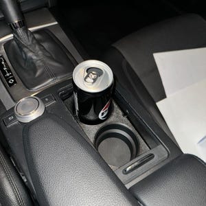 Może przedstawiać: Czarna i srebrna puszka po napoju gazowanym w uchwycie na kubek w samochodzie. Puszka ma czerwono-białe logo. Wnętrze samochodu jest czarne z fakturą przypominającą skórę.