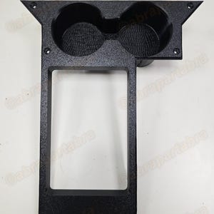 May include: Black plastic car console with two circular cup holders and a rectangular opening below. The console has a textured surface and mounting holes. The text "Copyright © 2025 abrapartabra ALL RIGHTS RESERVED" is visible.