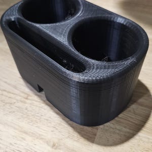 May include: A black 3D-printed car cup holder with two circular openings and a small rectangular slot. The cup holder is made of a textured plastic material and sits on a light-colored wooden surface.