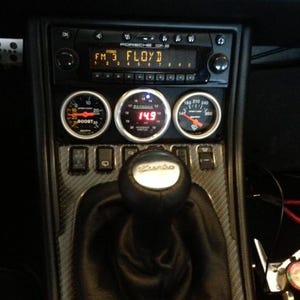 Puede incluir: Un interior de coche negro con un salpicadero de fibra de carbono, una radio que reproduce "FM 3 FLOYD" y una palanca de cambios con una perilla "Turbo". Hay tres indicadores, uno que marca "BOOST 30", otro que marca "149" y el tercero que marca "180 210 240 300".