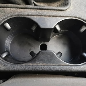 Op de afbeelding: Een zwarte plastic bekerhouder voor in de auto met twee ronde compartimenten en een klein centraal gat. De bekerhouder is geïntegreerd in een gestructureerd, donkergrijs oppervlak, waarschijnlijk de console van de auto.