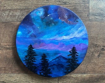 May include: A round painting depicting a night sky with a galaxy, stars, and a shooting star.  Silhouettes of trees and mountains are in the foreground.  The sky is painted in shades of blue, purple, and pink.