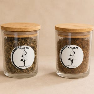 May include: Two clear glass jars with light brown bamboo lids, each filled with dried herbs. A circular white label on the front of each jar features a black line drawing of a person in a yoga tree pose, with the text KAIZEN Wellness Institute.