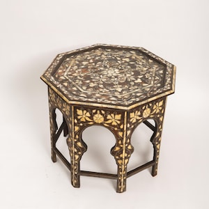 May include: An octagonal wooden side table with intricate floral inlay. The dark brown table features cream-coloured floral patterns on the tabletop and along the arched legs. The table is a decorative piece with a unique design.