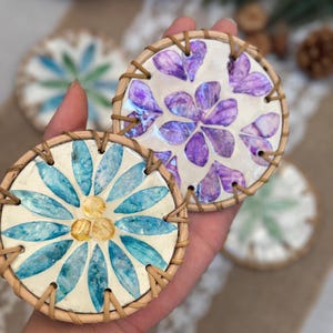 Puede incluir: Posavasos hechos a mano con diseños florales. Uno presenta una flor azul y amarilla, el otro una flor morada. Ambos tienen un fondo color crema y están enmarcados con ratán tejido. Decorativos y funcionales.