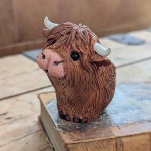 May include: A detailed figurine of a Highland cow, featuring brown fur, white horns, and a pink nose. The cow is positioned on top of a vintage book, showcasing a rustic aesthetic. The figurine's expressive eyes and textured details add to its charm.