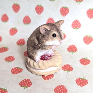 Op de afbeelding: Gedetailleerd beeldje van een kleine bruine muis die een rode aardbei vasthoudt op een met crème gevulde biscuit. De muis heeft zwarte ogen en staat tegen een achtergrond met aardbeipatronen.