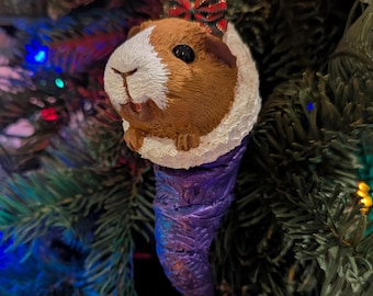 May include: A Christmas tree ornament featuring a guinea pig with brown and white fur, a red and black bow, and a purple, textured base. The ornament is hanging on a green Christmas tree.
