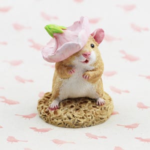 May include: A small, brown and white hamster figurine wearing a pink flower hat. The figurine is standing on a brown base with a textured surface.