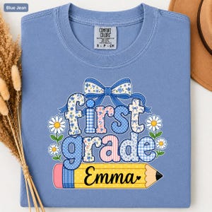 May include: A blue jean colored t-shirt featuring a graphic with the text first grade and the name Emma written on a yellow pencil. The design includes a blue patterned bow, white daisies, and various blue, pink, and white fabric patterns.