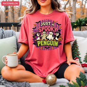 May include: A watermelon-colored t-shirt with a Halloween-themed graphic. The graphic features four penguins dressed as a witch, mummy, ghost, and a penguin with a witch hat holding a pumpkin. The text "JUST A SPOOKY PENGUIN" is displayed in purple and green.