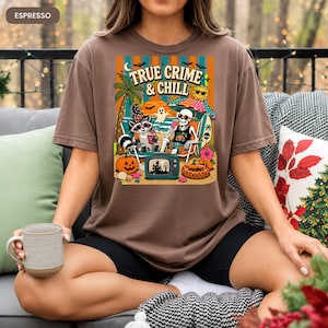 May include: Espresso-colored t-shirt with a colorful graphic that reads "TRUE CRIME & CHILL." The design features a raccoon and skeleton relaxing on a beach with a TV, palm trees, and pumpkins. The shirt is a comfortable fit.