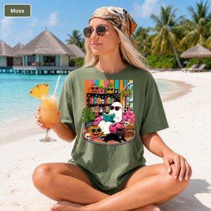 May include: Moss green t-shirt featuring a colorful graphic of a ghost reading a book, surrounded by Halloween-themed decorations, a black cat, and a flamingo. The shirt is worn by a person on a beach, holding a tropical drink.
