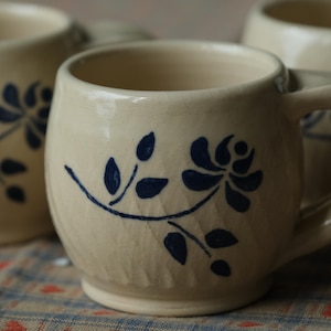 Può includere: Tazze in ceramica color crema con un design floreale blu. Le tazze hanno una forma arrotondata con un manico e sono decorate con un motivo di rose e foglie blu. Le tazze sono su una tovaglia fantasia.