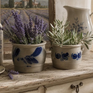 Puede incluir: Dos macetas de cerámica redondas de color gris claro con motivos florales pintados a mano en azul. Una contiene ramas de lavanda morada y la otra ramas de olivo verdes. Ambas tienen pequeñas asas laterales y un acabado esmaltado liso.