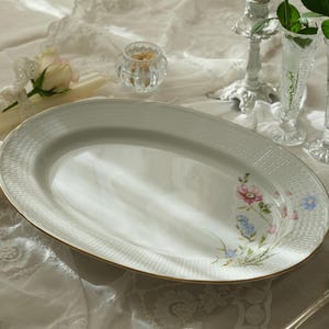 May include: An oval, white porcelain serving platter with a gold rim and floral accents. The platter is surrounded by crystal glassware, a silver spoon, and a white lace tablecloth. A rose and a napkin are also on the table.