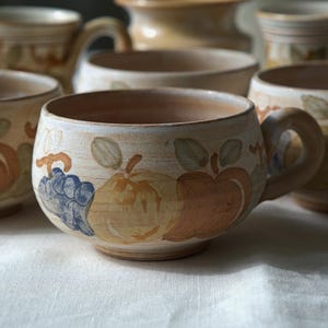 Può includere: Una collezione di tazze in ceramica con un motivo a frutta dipinto a mano. Ogni tazza presenta una base beige chiaro con illustrazioni di mele marroni, pere gialle e uva blu con foglie verdi. Le tazze hanno una forma arrotondata e un manico.