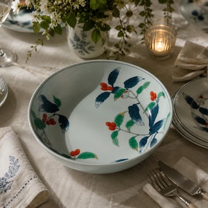 May include: A white ceramic serving bowl featuring a hand-painted botanical pattern of dark blue leaves, bright green foliage, and clusters of small red berries. The bowl has a smooth, rounded rim and a wide, shallow basin for serving food.