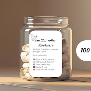 May include: A clear glass jar filled with rolled paper slips. The jar has a label with German text, including "Ein Glas voller Bibelverse" and "100 Bibelverse". The jar is on a wooden surface.